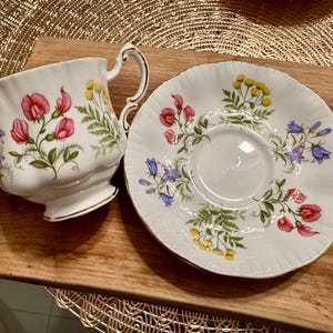 May include: A white teacup and saucer with a floral pattern featuring pink, blue, and yellow flowers. The floral pattern is on a white background.