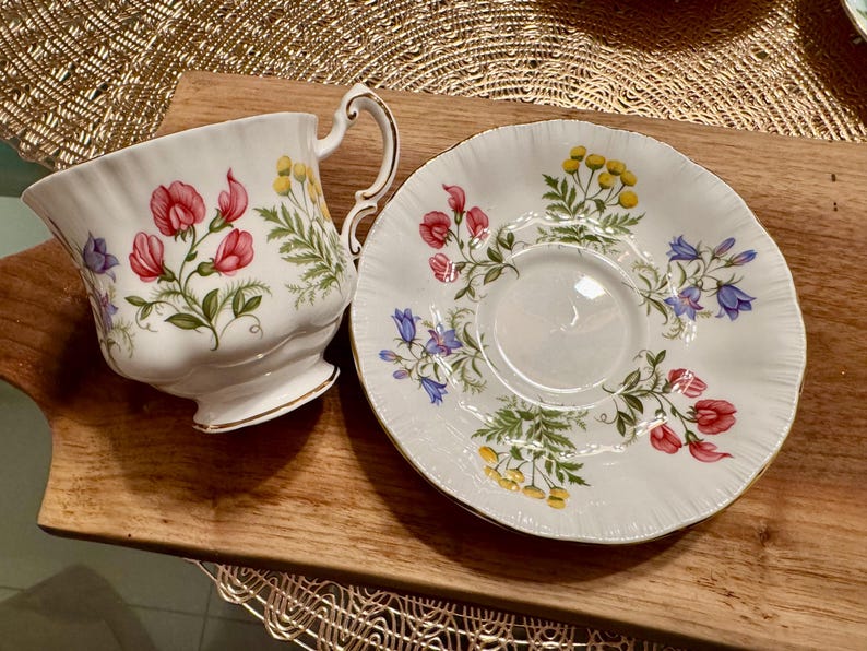May include: A white teacup and saucer with a floral pattern featuring pink, blue, and yellow flowers. The floral pattern is on a white background.
