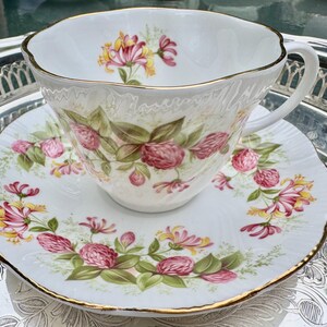 May include: A white teacup and saucer set with a floral design. The cup and saucer are decorated with pink and yellow flowers and green leaves. The rims are trimmed with gold. The set is placed on a silver tray.