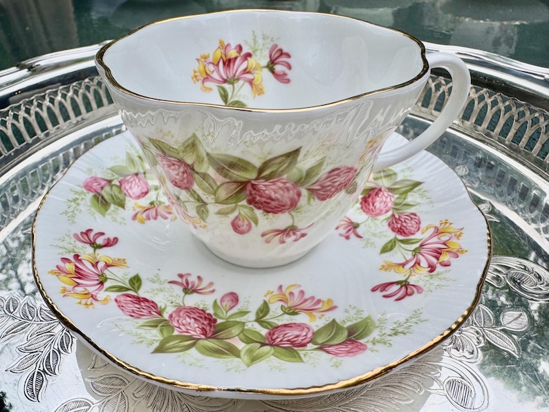 May include: A white teacup and saucer set with a floral design. The cup and saucer are decorated with pink and yellow flowers and green leaves. The rims are trimmed with gold. The set is placed on a silver tray.