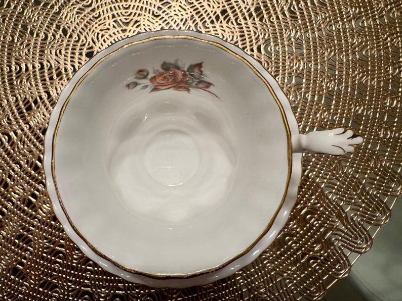 Pode incluir: Uma ch&aacute;vena de ch&aacute; branca com uma borda dourada e um desenho floral. A ch&aacute;vena de ch&aacute; tem uma pega com a forma de uma m&atilde;o.