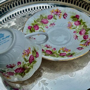 May include: White teacup and saucer set with a floral design in pink, yellow, and green. The teacup has a handle and is placed next to the saucer on a silver tray. The set is marked "Royal Heritage, Made in England."