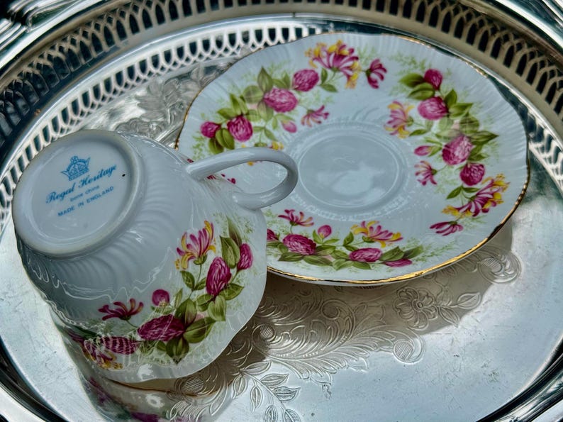 May include: White teacup and saucer set with a floral design in pink, yellow, and green. The teacup has a handle and is placed next to the saucer on a silver tray. The set is marked "Royal Heritage, Made in England."