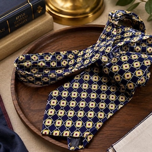 Puede incluir: Una corbata azul marino con un patr&oacute;n geom&eacute;trico repetitivo de diamantes amarillos y blancos con peque&ntilde;os centros rojos. La corbata est&aacute; doblada y descansa sobre una bandeja circular de madera, mostrando un acabado satinado y un extremo puntiagudo.