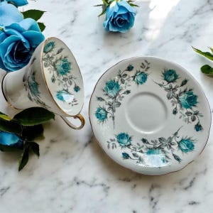 Peut inclure: Service &agrave; th&eacute; blanc avec une tasse et une soucoupe orn&eacute;es de roses turquoise et de bordures dor&eacute;es. La tasse est inclin&eacute;e, r&eacute;v&eacute;lant le motif int&eacute;rieur. La soucoupe est ronde avec un motif floral assorti. Des roses bleues sont dispos&eacute;es autour.