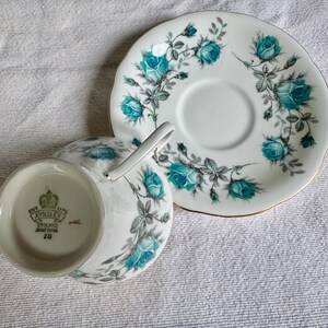 Peut inclure: Service &agrave; th&eacute; blanc avec une tasse et une soucoupe. Motif de roses turquoise et bordure dor&eacute;e. La tasse a une anse et la soucoupe un centre en creux. Le fond de la tasse porte la marque "AYNSLEY ENGLAND BONE CHINA 28".
