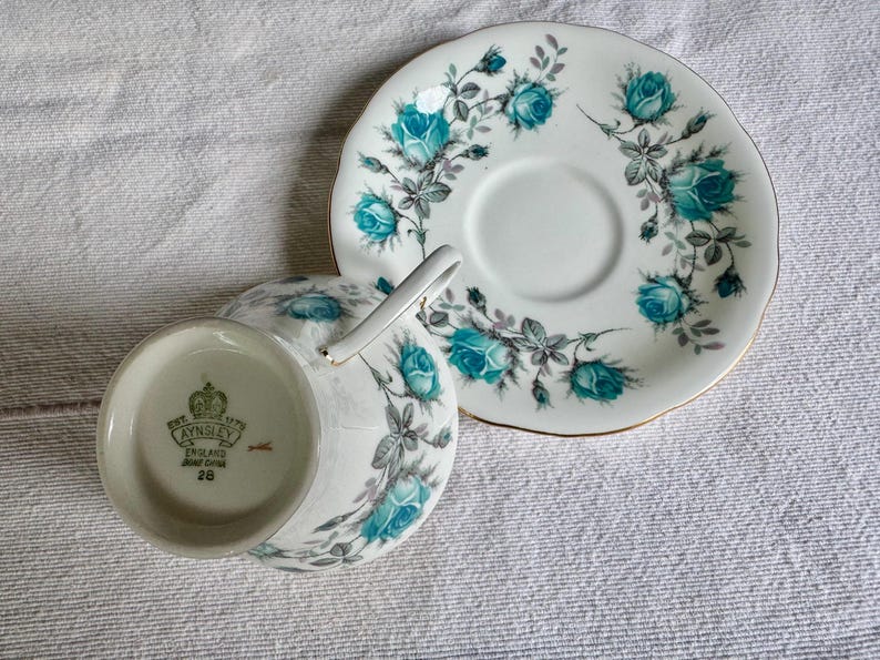 Peut inclure: Service &agrave; th&eacute; blanc avec une tasse et une soucoupe. Motif de roses turquoise et bordure dor&eacute;e. La tasse a une anse et la soucoupe un centre en creux. Le fond de la tasse porte la marque "AYNSLEY ENGLAND BONE CHINA 28".