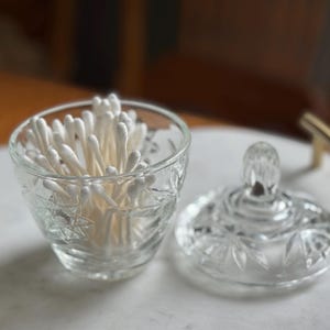 May include: A clear glass jar with a starburst pattern and a matching lid, filled with white cotton swabs. The jar features a faceted design and is positioned next to its removable lid, which has a small, rounded handle on top.