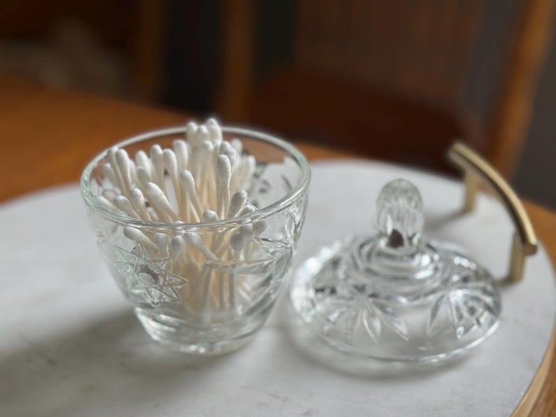 May include: A clear glass jar with a starburst pattern and a matching lid, filled with white cotton swabs. The jar features a faceted design and is positioned next to its removable lid, which has a small, rounded handle on top.