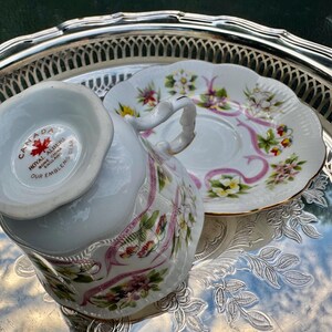 May include: A white teacup and saucer set with floral and pink ribbon designs, resting on a silver tray. The teacup has a curved handle and the saucer is scalloped. The teacup has the text "CANADA" and "ROYAL ALBERT BONE CHINA ENGLAND OUR EMBLEMS BEAR".