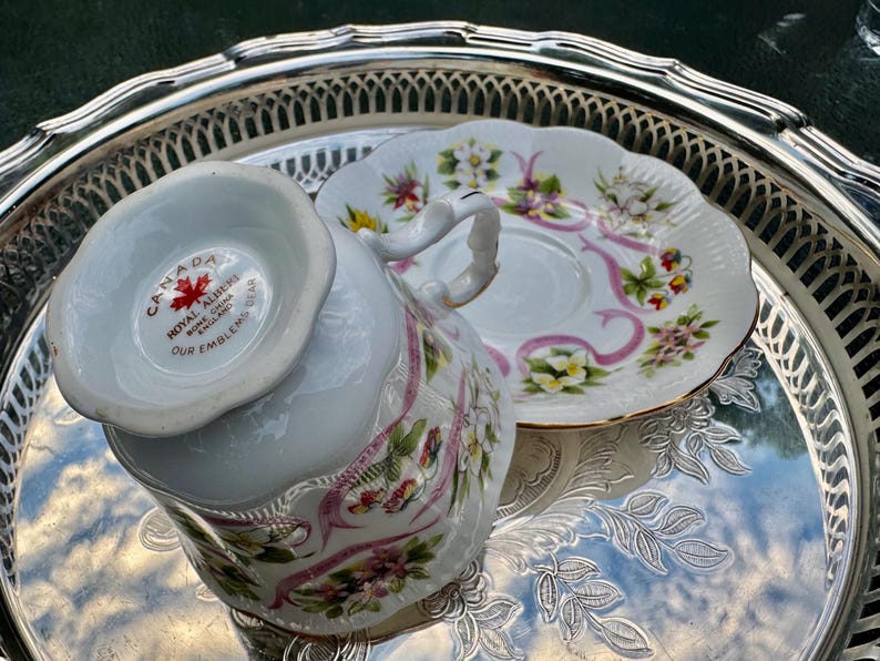 May include: A white teacup and saucer set with floral and pink ribbon designs, resting on a silver tray. The teacup has a curved handle and the saucer is scalloped. The teacup has the text "CANADA" and "ROYAL ALBERT BONE CHINA ENGLAND OUR EMBLEMS BEAR".