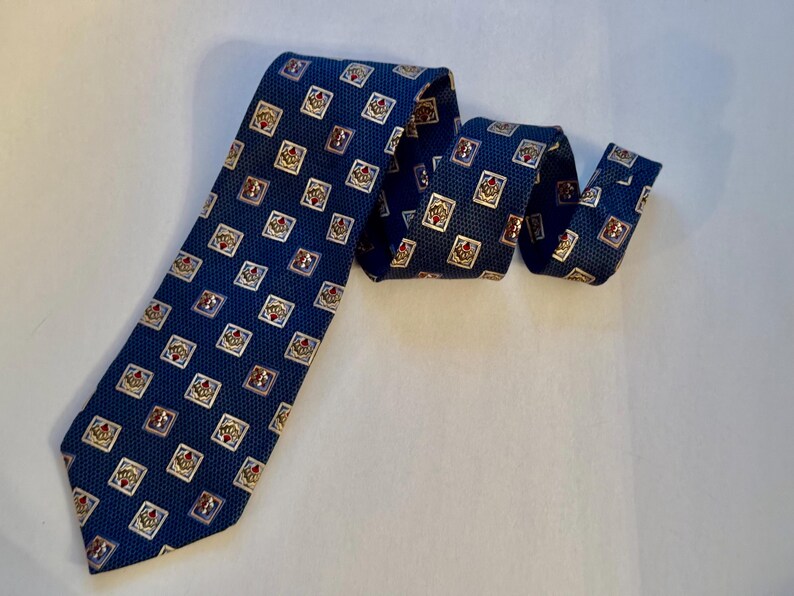 May include: A dark blue necktie featuring a repeating pattern of small, light-colored squares. Each square contains a detailed, centered graphic element with red and gold accents. The tie has a textured, woven appearance throughout the fabric.