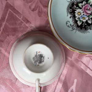 May include: A Paragon fine bone china teacup and saucer set. The saucer features a pale blue rim, a gold edge, and a central black medallion with pink and white floral motifs. The teacup base shows the Paragon maker's mark and royal crest.
