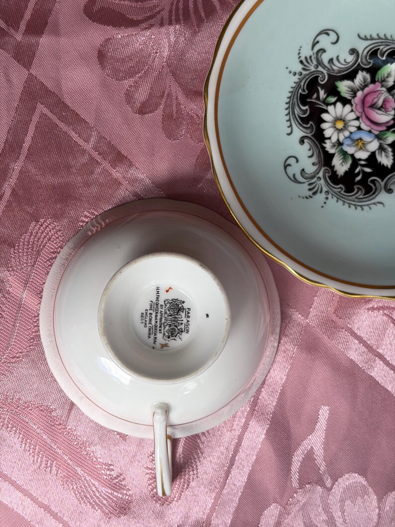 May include: A Paragon fine bone china teacup and saucer set. The saucer features a pale blue rim, a gold edge, and a central black medallion with pink and white floral motifs. The teacup base shows the Paragon maker's mark and royal crest.