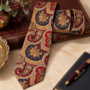 May include: A necktie featuring a detailed paisley pattern in shades of deep red, navy blue, and golden tan. The fabric displays intricate swirling motifs and floral-inspired shapes arranged in a repeating design across the entire length.