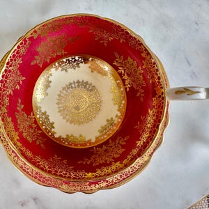 May include: A vintage teacup and saucer set. The teacup and saucer are a vibrant red with intricate gold floral patterns and trim. The teacup handle is white with gold accents. The small saucer in the center is cream-colored with a gold design.