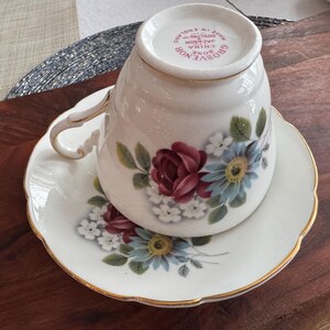 May include: A white teacup and saucer with a floral design. The teacup has a gold rim and the saucer has a scalloped edge. The teacup has the words "Grosvenor China, Jackson, Made in England" on the bottom.