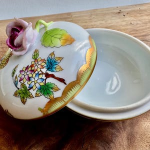 May include: A white porcelain trinket box with a floral design. The lid features a pink rose with green leaves and a gold trim. The box is decorated with colorful flowers and leaves.