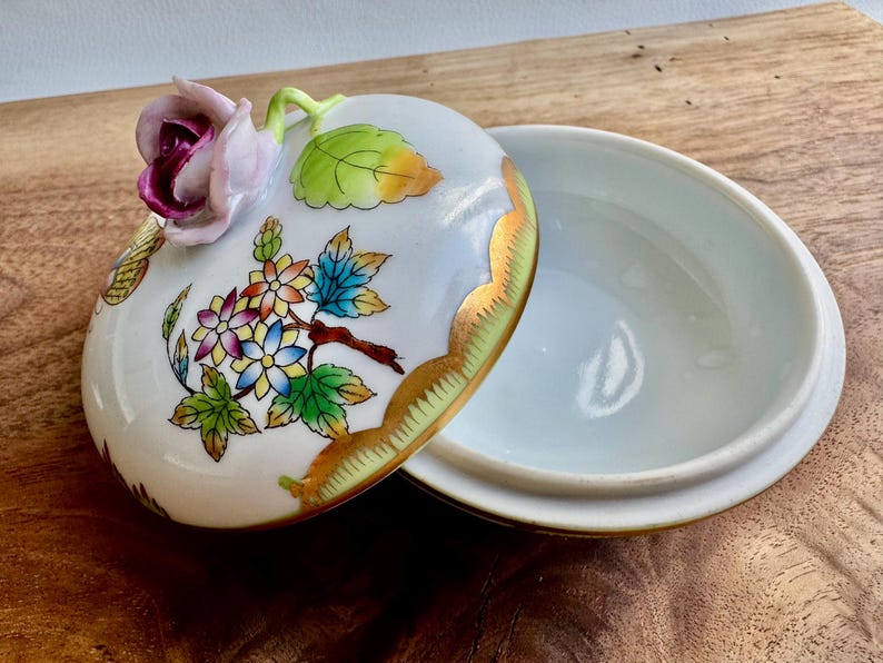 May include: A white porcelain trinket box with a floral design. The lid features a pink rose with green leaves and a gold trim. The box is decorated with colorful flowers and leaves.