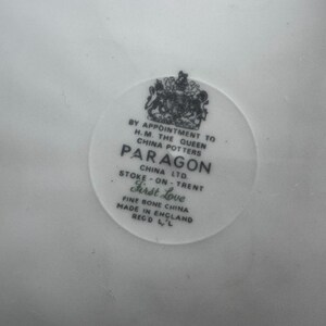 May include: A white porcelain plate with a black and white logo for Paragon China Ltd. The logo includes the text "By Appointment to H.M. The Queen China Potters Paragon China Ltd. Stoke-on-Trent First Love Fine Bone China Made in England Reg'd L'L."
