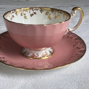Peut inclure: Service &agrave; th&eacute; rose compos&eacute; d'une tasse et d'une soucoupe. La tasse a un bord festonn&eacute; et une anse dor&eacute;e. La soucoupe et le bord de la tasse sont d&eacute;cor&eacute;s de motifs floraux dor&eacute;s. L'int&eacute;rieur de la tasse est blanc avec des accents floraux dor&eacute;s.