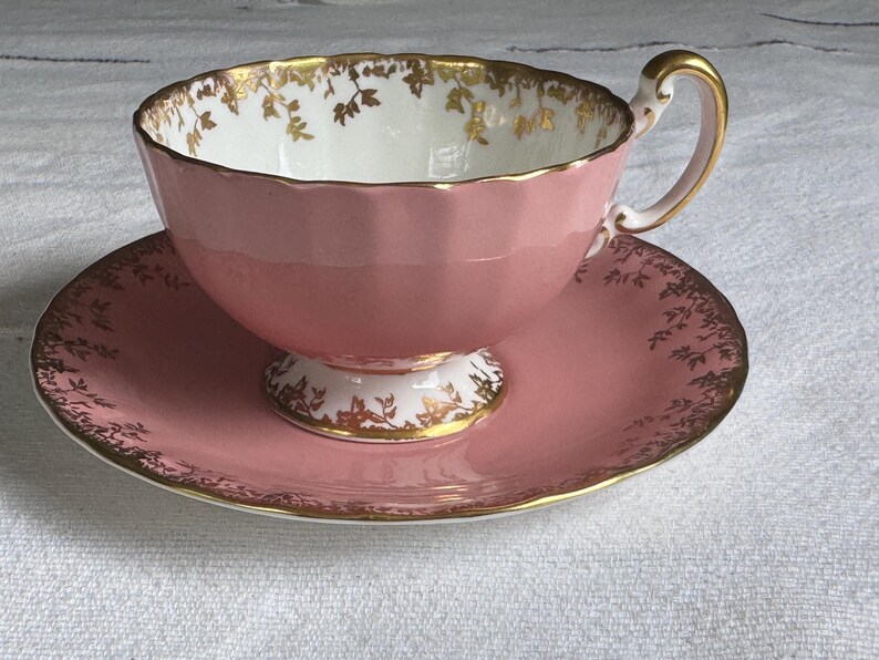 Peut inclure: Service &agrave; th&eacute; rose compos&eacute; d'une tasse et d'une soucoupe. La tasse a un bord festonn&eacute; et une anse dor&eacute;e. La soucoupe et le bord de la tasse sont d&eacute;cor&eacute;s de motifs floraux dor&eacute;s. L'int&eacute;rieur de la tasse est blanc avec des accents floraux dor&eacute;s.