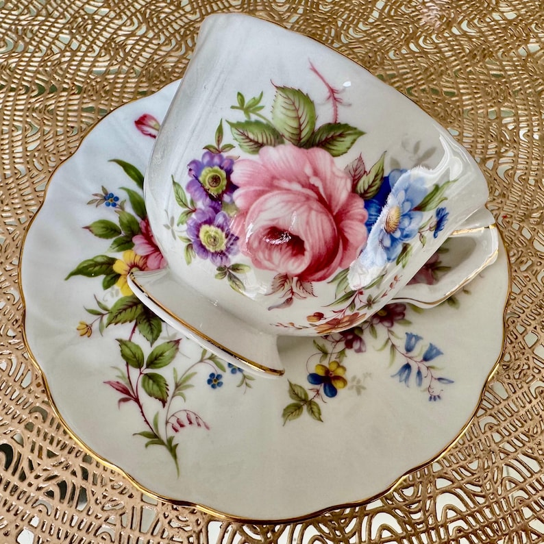 May include: A white teacup and saucer with a floral pattern. The cup and saucer have a gold trim. The floral pattern features pink, purple, blue, and yellow flowers.