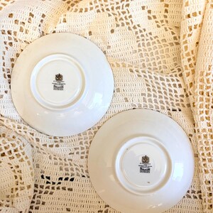 May include: Two white ceramic saucers with a circular base and a central stamp. The stamp features a black crest and the text BY APPOINTMENT TO HER MAJESTY THE QUEEN PARAGON FINE BONE CHINA ENGLAND REGD. The saucers are smooth and circular.
