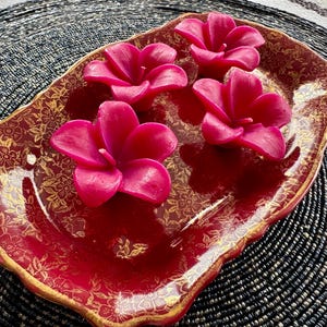 May include: Four pink flower-shaped candles on a red and gold decorative tray. The tray has a scalloped edge and a floral pattern.