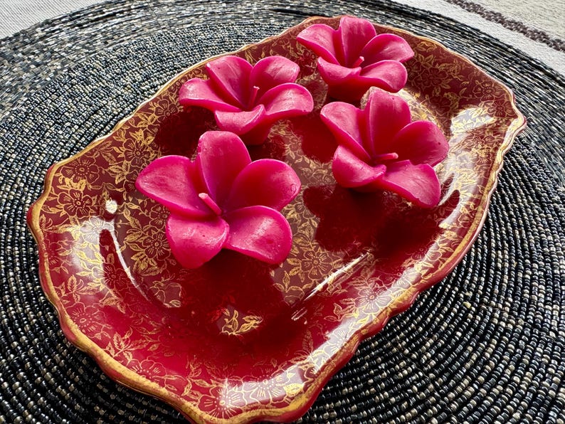 May include: Four pink flower-shaped candles on a red and gold decorative tray. The tray has a scalloped edge and a floral pattern.