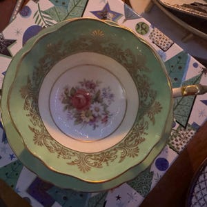 Peut inclure: Une tasse &agrave; th&eacute; et sa soucoupe vert clair avec un bord festonn&eacute;, des d&eacute;tails en filigrane dor&eacute; et un bouquet floral central compos&eacute; de fleurs roses, blanches et violettes. La tasse poss&egrave;de une anse dor&eacute;e et une base int&eacute;rieure blanche.