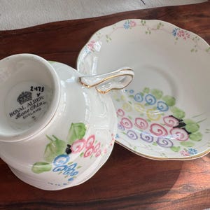 May include: A vintage Royal Albert Bone China teacup and saucer set with a floral design. The cup features a pink, blue, and green floral pattern with a gold trim. The saucer has a similar floral pattern with a gold rim.