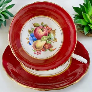May include: A red teacup and saucer set with a gold rim and a white floral design featuring fruit. The cup has a scalloped edge.