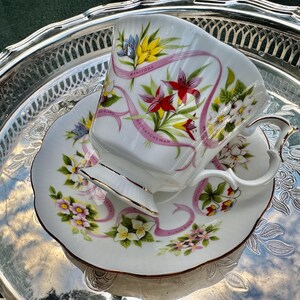 May include: A white teacup and saucer set with floral and ribbon designs. The cup and saucer feature colorful flowers and pink ribbons with the words "Manitoba" and "Saskatchewan". The set is placed on a silver tray.