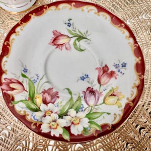 May include: A white porcelain saucer with a red border and floral design. The saucer features a central well and is decorated with red, purple, and yellow tulips, white flowers, and green leaves. The rim has a scalloped edge.