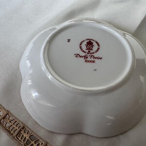 May include: A white, scalloped-edge dish with the text "Derby Posies" and "English Bone China" in red. The dish has a floral design and is likely vintage. The dish is on a white surface.