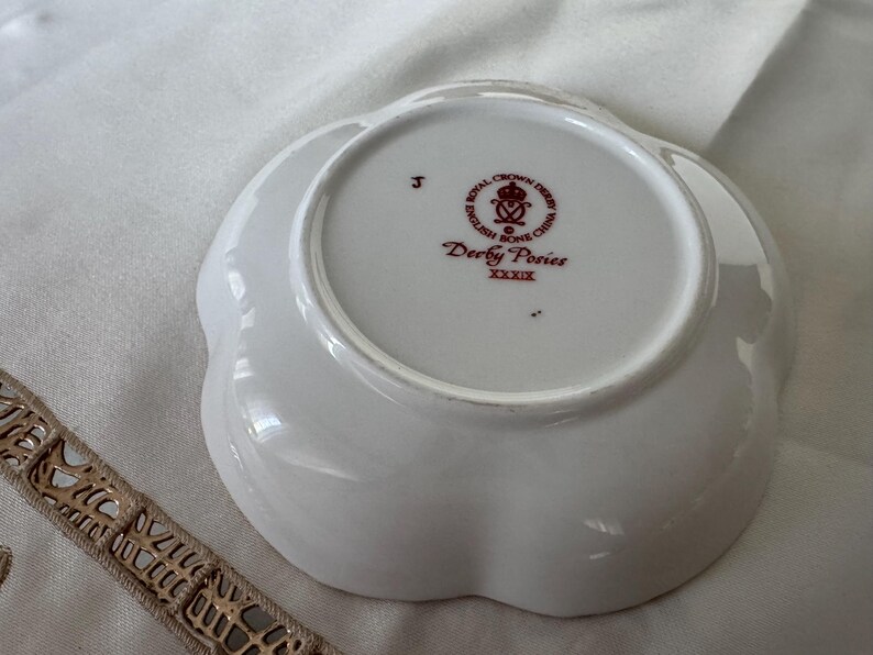 May include: A white, scalloped-edge dish with the text "Derby Posies" and "English Bone China" in red. The dish has a floral design and is likely vintage. The dish is on a white surface.