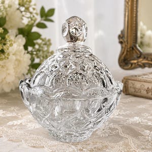 May include: A clear glass candy dish with a matching lid, featuring a faceted diamond-cut pattern across the surface. The lid is topped with a rounded, textured finial, and the bowl has a scalloped rim, creating a reflective, multi-faceted appearance.