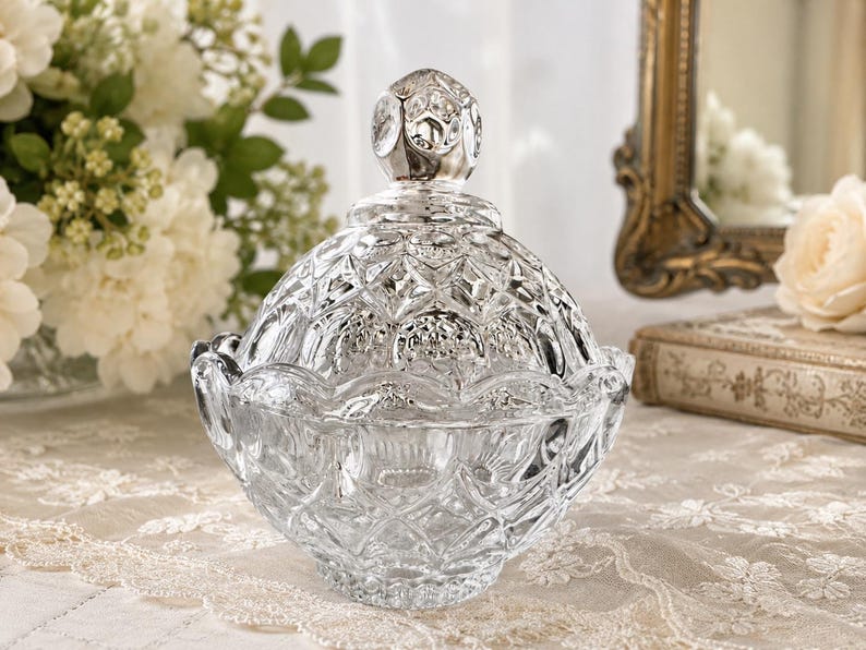 May include: A clear glass candy dish with a matching lid, featuring a faceted diamond-cut pattern across the surface. The lid is topped with a rounded, textured finial, and the bowl has a scalloped rim, creating a reflective, multi-faceted appearance.