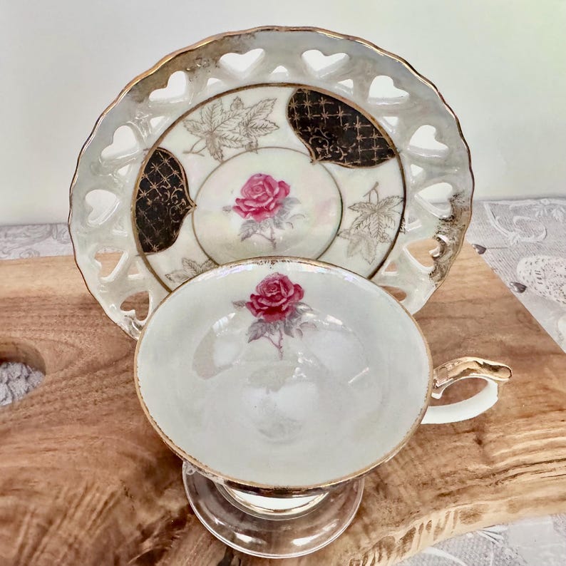 Puede incluir: Un juego de taza y platillo de t&eacute; vintage. La taza y el platillo blancos presentan un dise&ntilde;o de rosa rosa. El platillo tiene un borde en forma de coraz&oacute;n y ribete dorado. La taza tiene un asa dorada y se asienta sobre una base de vidrio transparente. El juego est&aacute; sobre una superficie de madera.