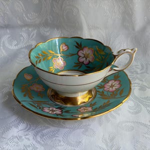 May include: A porcelain teacup and saucer set featuring a vibrant turquoise background with hand-painted pink flowers and gold leaf foliage accents. The cup has a scalloped rim and a gold-trimmed base, while the saucer matches the floral design.