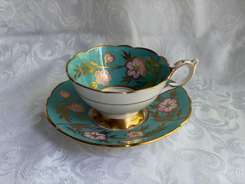 May include: A porcelain teacup and saucer set featuring a vibrant turquoise background with hand-painted pink flowers and gold leaf foliage accents. The cup has a scalloped rim and a gold-trimmed base, while the saucer matches the floral design.