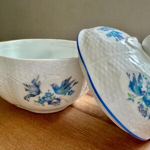 May include: White ceramic trinket dish with a lid, featuring blue floral and bird designs. The dish has a textured surface and a blue rim. The lid has a tulip-shaped handle. A decorative piece for storing small items.