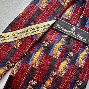 May include: An Ermenegildo Zegna necktie featuring a repeating pattern of golf bags on a background of alternating maroon textured stripes and black stripes with blue accents. The tie includes a branded label and a cream-colored interior fabric tag.