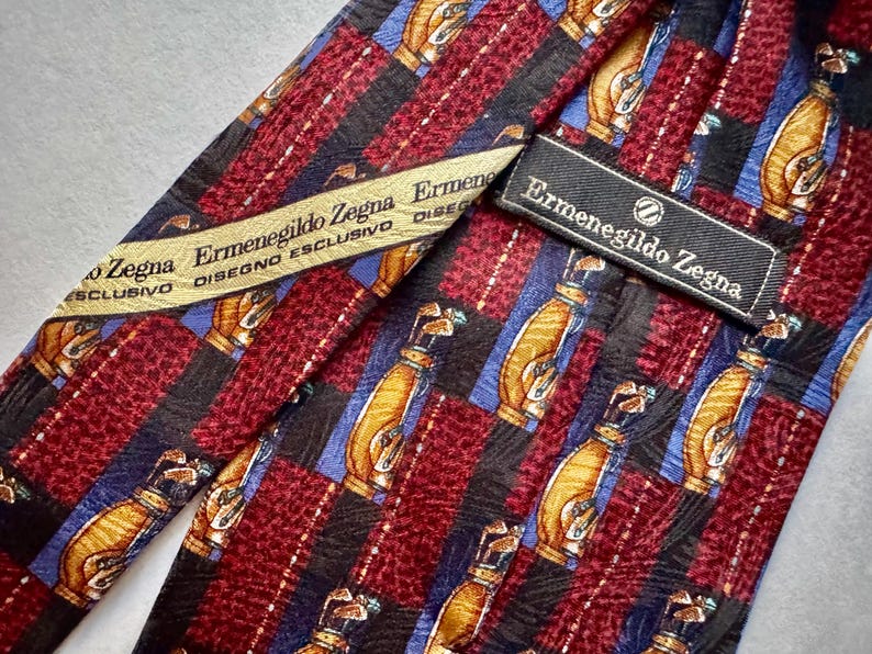 May include: An Ermenegildo Zegna necktie featuring a repeating pattern of golf bags on a background of alternating maroon textured stripes and black stripes with blue accents. The tie includes a branded label and a cream-colored interior fabric tag.
