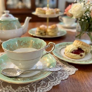 Peut inclure: Un ensemble tasse et soucoupe en porcelaine avec une bordure vert menthe p&acirc;le et des motifs en filigrane dor&eacute;. Une cuill&egrave;re en argent avec un motif floral en relief repose sur la soucoupe. Une th&eacute;i&egrave;re assortie, des scones et des roses sont visibles.