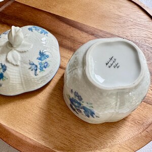 May include: White ceramic lidded dish with blue floral designs. The lid features a rose-shaped handle. The dish is open, revealing the interior. The dish and lid are on a wooden tray.