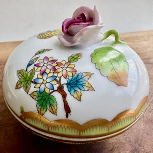 May include: A white porcelain trinket box with a floral design. The lid features a 3D pink rose with green leaves. The box has a gold rim and a green and gold base.