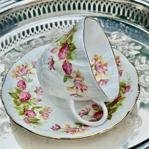 May include: A white teacup and saucer set with a floral design featuring pink and yellow flowers and green leaves. The teacup is resting on the saucer, which has a gold trim. The set is on a silver tray.