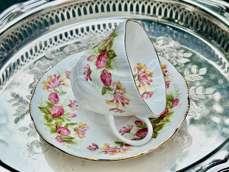 May include: A white teacup and saucer set with a floral design featuring pink and yellow flowers and green leaves. The teacup is resting on the saucer, which has a gold trim. The set is on a silver tray.